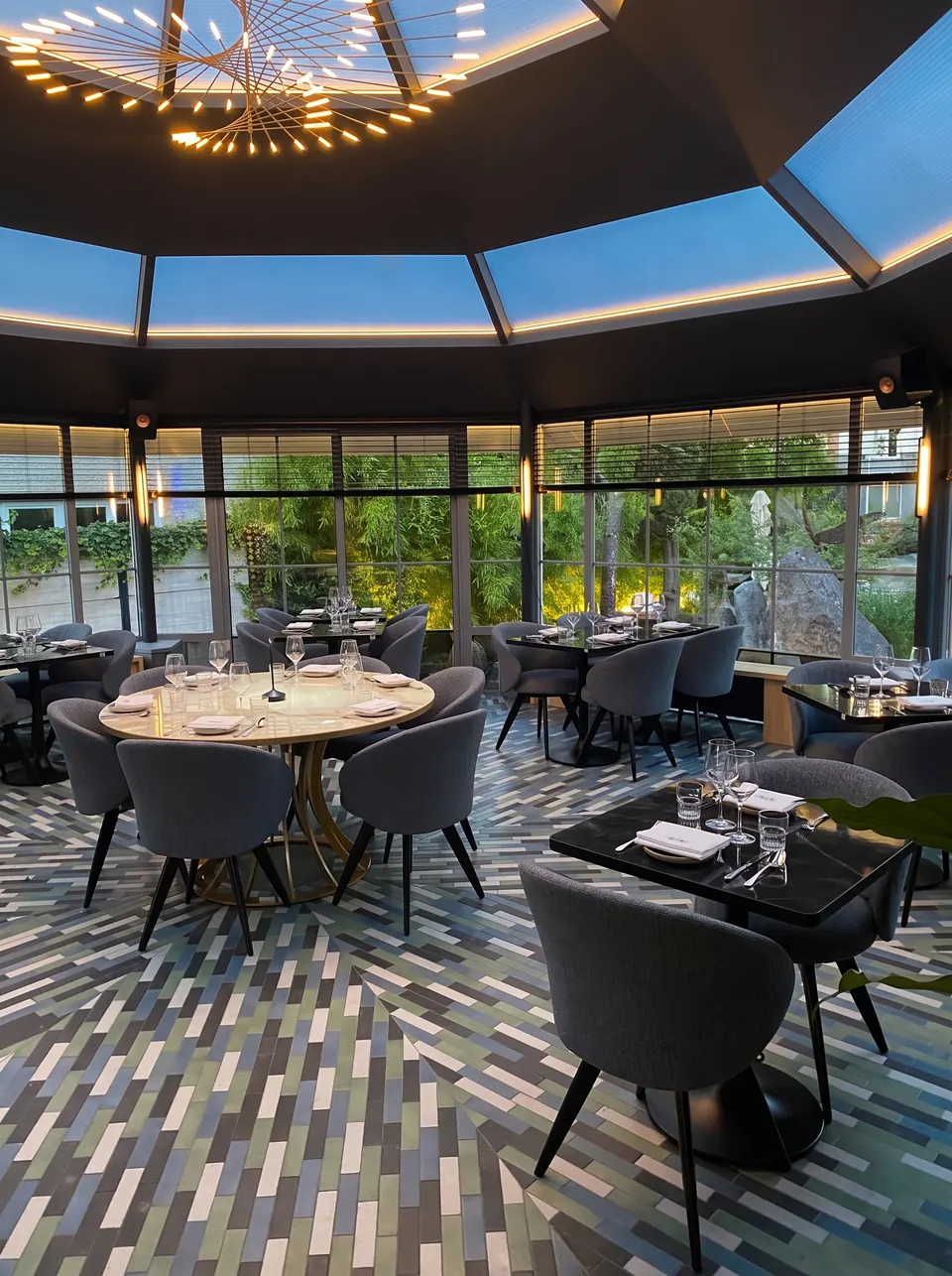 La salle de la verrière le soir, sous le lustre en soleil, les tables dressées